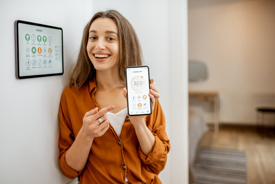 Portrait of a young and happy woman near a touch screen panel for managing smart home. Holding smart phone with a simlpe and convinient mobile application for smart home