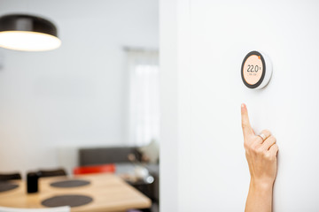 Round smart thermostat with touch screen installed on the wall with a hand trying to reach it. Smart home heating regulation concept