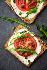 Healthy Breakfast Idea. Tomato sandwich with cream cheese and fresh rocket on black stone background. Top view. 