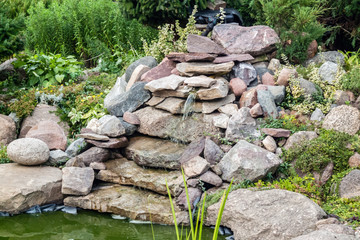 a small spring flowing from a pile of stones in the garden next to a stream and lush vegetation zoom on full frame