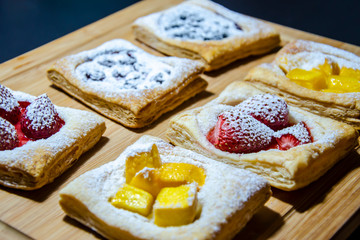 Homemade puff pastry with red strawberries and mangoes sprinkled with white icing sugar on a beige wooden board