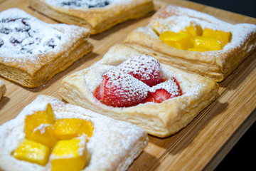Homemade puff pastry with red strawberries and mangoes sprinkled with white icing sugar on a beige wooden board