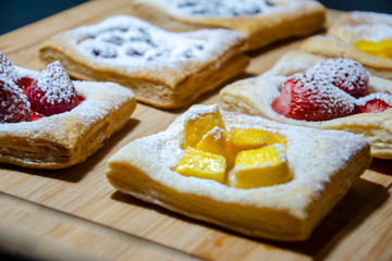 Homemade puff pastry with red strawberries and mangoes sprinkled with white icing sugar on a beige wooden board