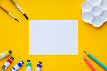 top view of brushes, color tubes, pencils, palette and white paper on yellow background, copy...