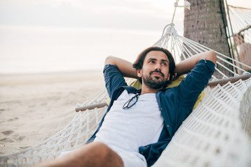 A man enjoys calm, lies in a hammock on the background of the ocean and sunset.