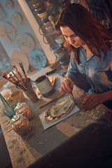 Talented young woman is working on new decorative plate at her cozy workshop.