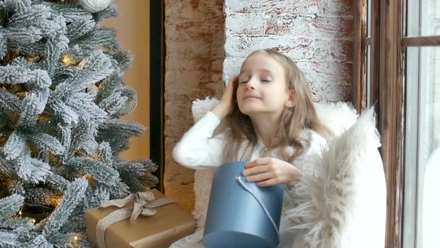Cute blond girl is opening dark blue gift box she recieved sitting near christmas tree, happy childhood and xmas celebration concepts