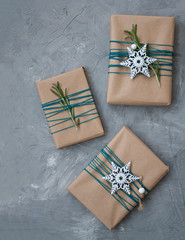Three Christmas gifts in craft paper wrapped in blue thread, with white snowflakes and rosemary branches on a gray concrete background. There is a place for a welcome text..