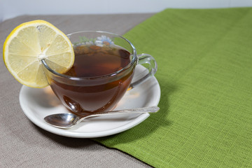On a white saucer a cup of tea with lemon and a silver spoon. Under the cup, two wipes are green and gray.