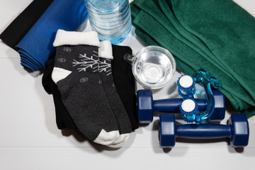 On a white background, items for fitness - a folded green towel, socks, a sports swimsuit, dumbbells, a hand massager, a rug, a bottle and a cup of water.