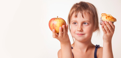 Studio shot of сute little girl holding apple in one hand and cake in another. Child trying to make decision between healthy and unhealthy food. Selective focus on apple and cake.