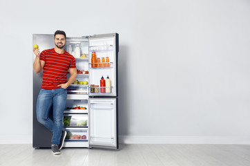 Happy young man with apple near open refrigerator indoors, space for text