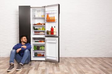 Happy young man sitting on floor near refrigerator indoors, space for text