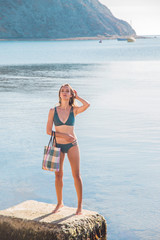 Millennial blonde woman with a beach bag in her hands by the sea in travel and relaxation.