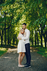 A beautiful and gentle girl with light hair and a white dress is walking in a sunny summer forest with her handsome guy in a white shirt and dark pants