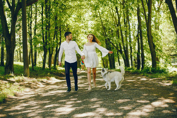 Fototapeta premium A beautiful and gentle girl with light hair and a white dress is walking in a sunny summer forest with her handsome guy in a white shirt and dark pants and they playing with cute gray dog