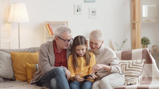 Knee-up shot of young Caucasian teenage girl sitting on couch with her loving grandparents, taking selfie together on smartphone, then admiring photo on screen and smiling