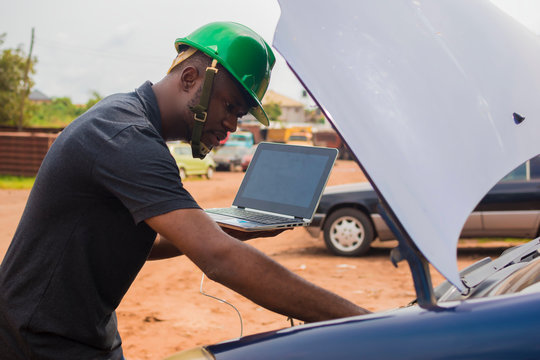 Young black engineer making a phone call and also holding his laptop checking out something with a broken down car behind him