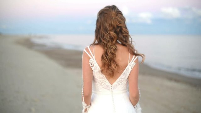 Back of a beautiful bride in a wedding dress walking on the seashore on background of the rose sunset cloud