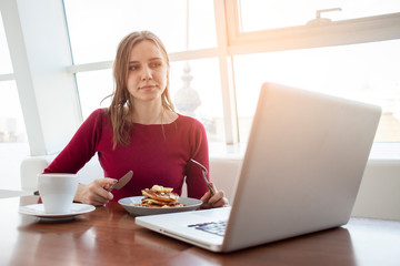 young girl freelancer sits in a cafe with a laptop, drinks coffee and has breakfast, a student eats in the morning with a computer