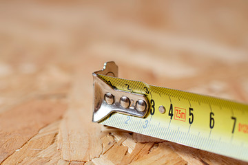 A tip of yellow metal tape measure lying on chip board. Blurred background.
