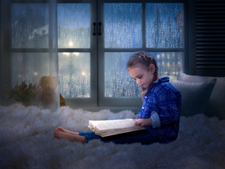 Little girl with a book late at night in a cozy children's room on the bed