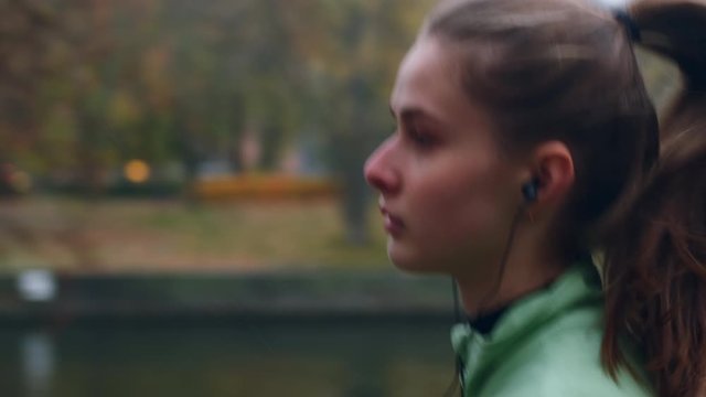 Tracking shot of attractive sporty girl in earphones dreamily running in autumn city park