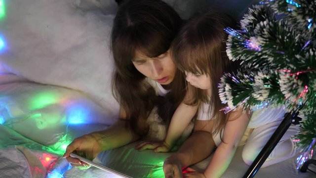 child and mother are playing with tablet computer in room. concept o happy childhood and family. Mom and daughter on Christmas evening watching cartoons on a tablet in children's room with garlands.