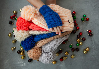 Obraz na płótnie Canvas Hands of young beautiful woman in blue mittens holding many colored bright warm winter hats. On a gray background are bright Christmas decorations.