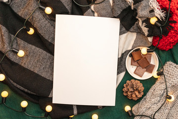 Christmas mockup, with a postcard, wishes, letter on the blanket with a kettle, leaf, tea, cone, chocolate and xmas tree lights.  Background top view blank space for text.