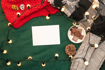 Christmas mockup, with a postcard, wishes, letter on the blanket with a kettle, leaf, tea, cone, chocolate and xmas tree lights.  Background top view blank space for text.
