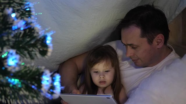 concept of family Christmas. child and dad on Christmas evening play on tablet, in children's room, in tent with garlands. baby and father are playing in room. concept of happy childhood and family.