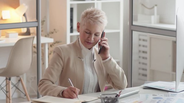 Waist-up shot of smiling mature Caucasian woman in smart business suit, with short grey hair sitting in office, talking on mobile phone, listening and writing down information in journal