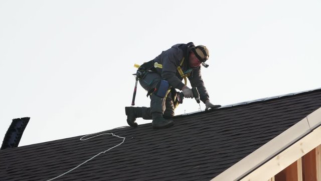 Professional builder in safety harness is making roof ridge using hydraulic hammer. Works at height. Frame house under construction