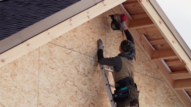 Builder in gloves is covering the wooden frame with plywood using hydraulic hammer and standing on ladder. Frame house under construction