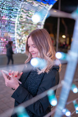 Young beautiful caucasian girl looking and touching the smartphone on the street with Christmas lights in the background