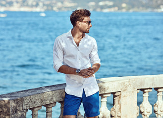 Portrait of handsome man wearing elegant white shirt and sunglasses, standing near the lake