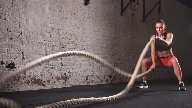 Athletic woman exercises with battle ropes as part of her fitness gym workout. Slow motion