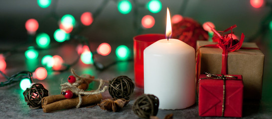 Red and white festive candles burn with a yellow flame on a table among gifts in craft paper, rattan balls, cinnamon sticks and anise stars on the background of a multi-colored Christmas garland