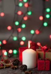 Obraz na płótnie Canvas Red and white festive candles burn with a yellow flame on a table among gifts in craft paper, rattan balls, cinnamon sticks and anise stars on the background of a multi-colored Christmas garland