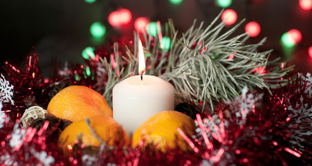Fototapeta na wymiar A white candle burns with flame on a table among gifts in craft paper, rattan balls, spruce wreath, tangerines, cinnamon sticks on a background of multi-colored Christmas garland