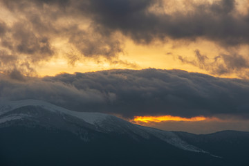 Fototapeta premium Evening dramatic sky with colorful burning clouds against the backdrop of mountain houses in winter