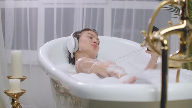 A young woman lies in the bath wearing headphones and looking at the screen of a smartphone
