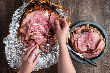 Woman’s hand pulling pieces of ham off a Spiral cut glazed and cooked ham in a foil wrapper on a...