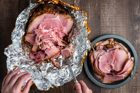Woman’s hand cutting pieces of ham off a spiral cut glazed and cooked ham in a foil wrapper on a wood table, black plate with slices