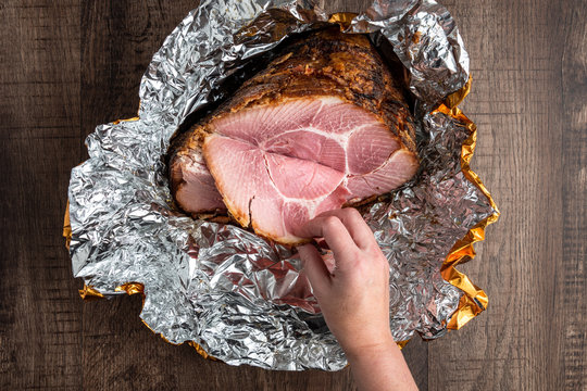 Woman’s hand pulling a piece of ham off a spiral cut glazed and cooked ham in a foil wrapper on a wood table