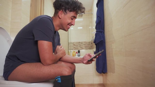 Young man sitting on the toilet, using the cell phone while laughing