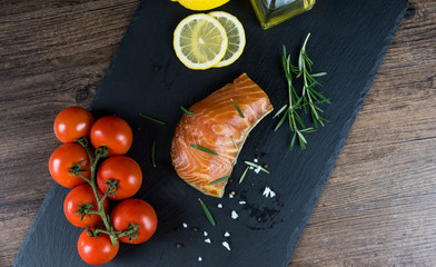 Grilled salmon steak on vintage wooden board with fork, olive oil, vegetables, herbs and spices on dark concrete background. Raw salmon fillet with vegetables, spices and lemon on wooden table.
