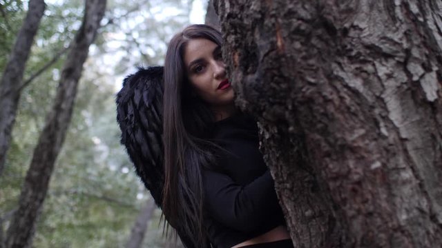 Beautiful gothic model with black angel wings peaks out from behind a tree in the forest.