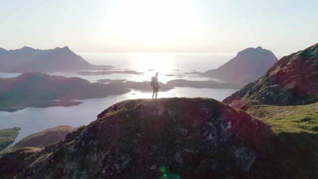 4K slow motion drone shot with orbit motion of young Norwegian climber with backpack standing on top of mountain admiring the panoramic sunset view in Lofoten Vester&aring;len in Norway.